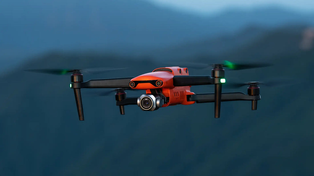 A vibrant orange drone with a camera hovers over a blurred mountain landscape. The sky is overcast, and lights on its arms are green, indicating readiness.