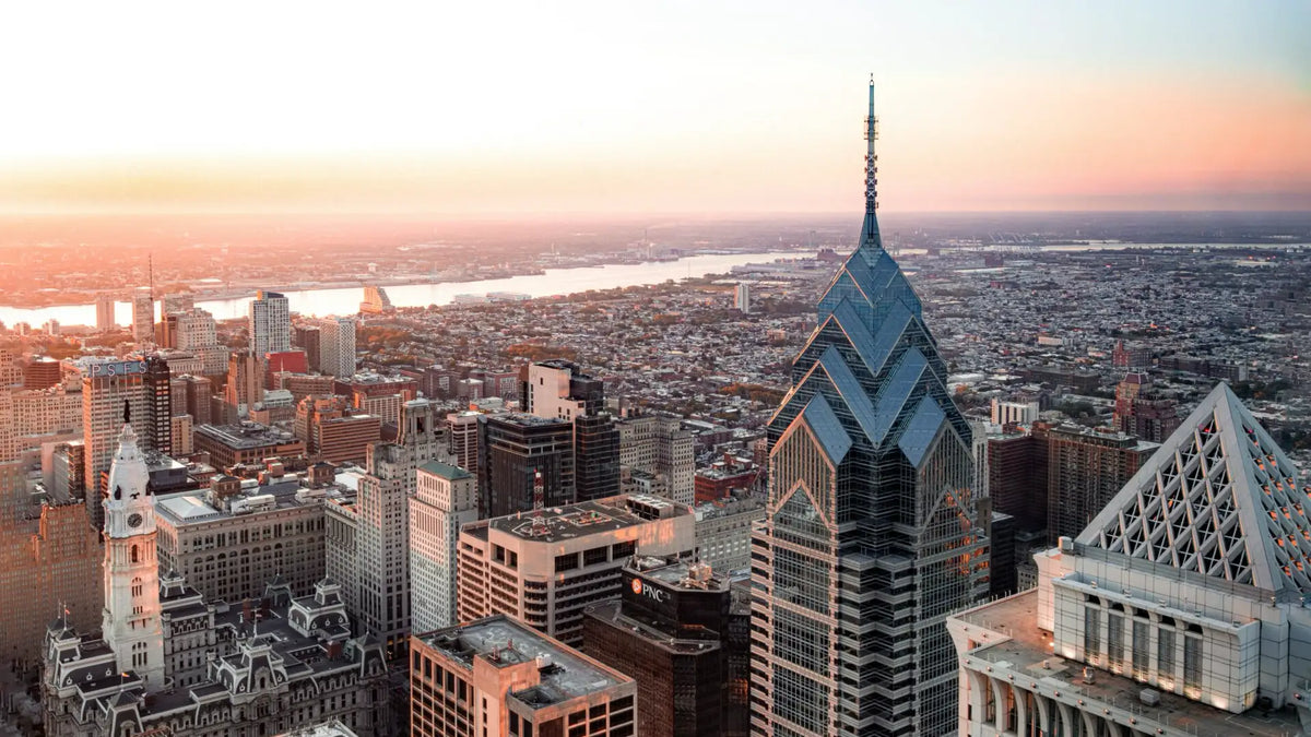 Aerial view of a cityscape at sunset with prominent tall buildings, including a glass skyscraper with a spire, and an expansive river in the background.