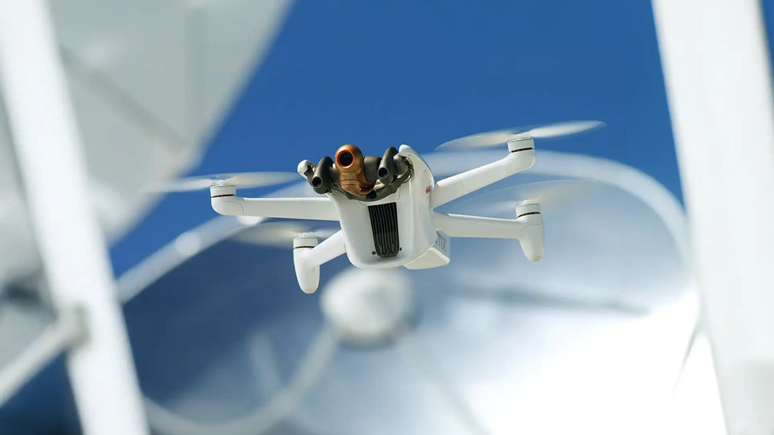 A small white drone with four propellers hovers, set against a clear blue sky. It carries a toy figurine, adding a playful touch to the tech-focused scene.
