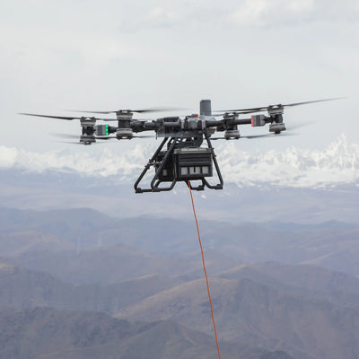 DJI Dual-Battery Lifting System in flight beneath FlyCart 100, lowering a tether over mountains